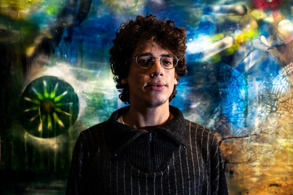 A young man with curly medium length hair and glasses against a multicoloured wall 