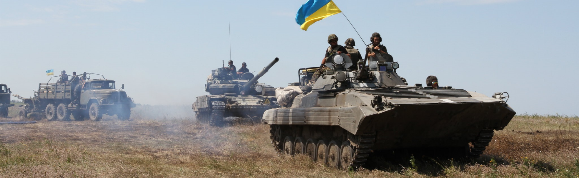 Two Ukrainian battle tanks and a truck on the move through a field.